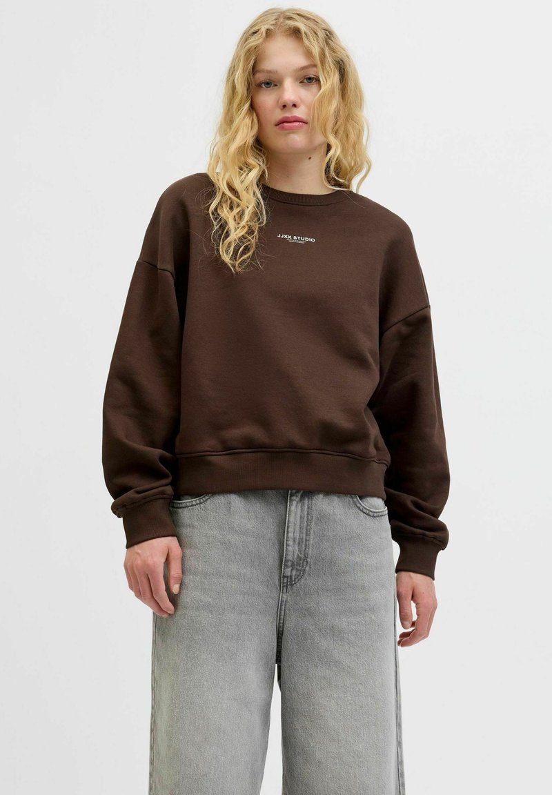 Bruine, oversized sweatshirt met ronde hals en ribgebreide boorden; voorzien van witte tekstlogo "JJXX STUDIO" op de voorkant. Draagt bij grijze jeans.