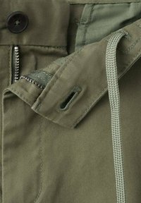 Zbliżenie zielonych spodni pokazujące guziki, metalowy zamek błyskawiczny, sznurek ściągający oraz oczka na guziki na materiale.