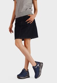 Donna con un top senza maniche grigio, una gonna di jeans scura e sneakers con zeppa blu navy, in piedi con le gambe incrociate su uno sfondo bianco.