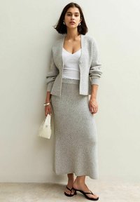 Un cardigan en maille gris clair sur un débardeur blanc, associé à une jupe moulante en maille gris et des sandales noires. Tient un petit sac blanc.