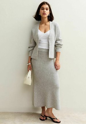 Light gray knitted cardigan over a white tank top, paired with a fitted gray ribbed skirt and black sandals. Holds a small white bag.