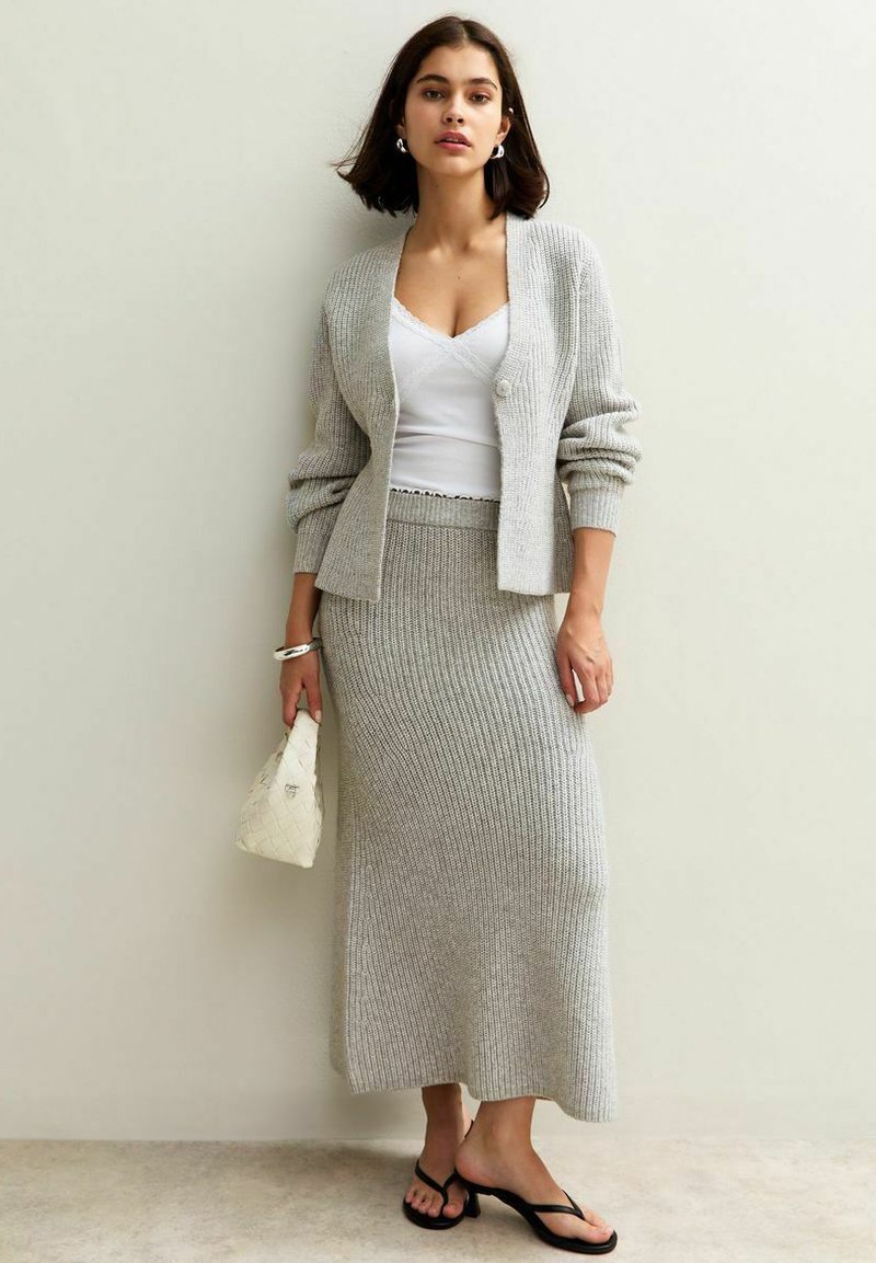Un cardigan en maille gris clair sur un débardeur blanc, associé à une jupe moulante en maille gris et des sandales noires. Tient un petit sac blanc.