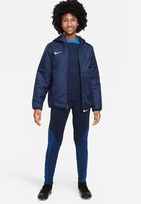 Marineblaues Sportjacke mit Kapuze, Reißverschluss und Logo; kombiniert mit marineblauen Leggings mit blauem geometrischem Muster. Sportschuhe vervollständigen das Outfit.