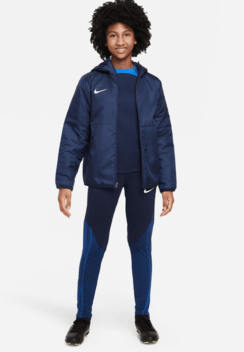 Marineblaues Sportjacke mit Kapuze, Reißverschluss und Logo; kombiniert mit marineblauen Leggings mit blauem geometrischem Muster. Sportschuhe vervollständigen das Outfit.