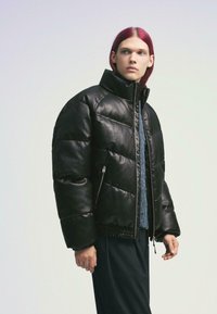 Doudoune en cuir noir avec un col montant, dotée de poches zippées, de poignets élastiqués et d'une finition brillante. Le modèle porte un pull bleu en dessous.