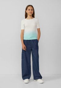 T-Shirt mit weißem und türkisfarbenem Farbverlauf und kurzen Ärmeln, kombiniert mit weit geschnittenen Denim-Hosen mit seitlichen Taschen, getragen mit weißen Sneakers.