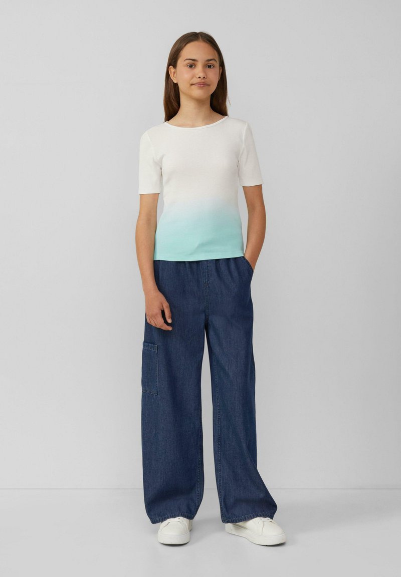 T-Shirt mit weißem und türkisfarbenem Farbverlauf und kurzen Ärmeln, kombiniert mit weit geschnittenen Denim-Hosen mit seitlichen Taschen, getragen mit weißen Sneakers.