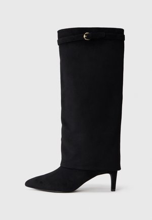Bota de camurça preta até ao joelho com bico pontiagudo, saltinho fino e uma fita decorativa com uma fivela circular na parte superior.