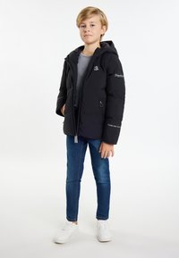 Svart puffjacka med dragkedja framtill, huva och broderade detaljer. Bärs över en grå skjorta, tillsammans med blå jeans och vita sneakers.