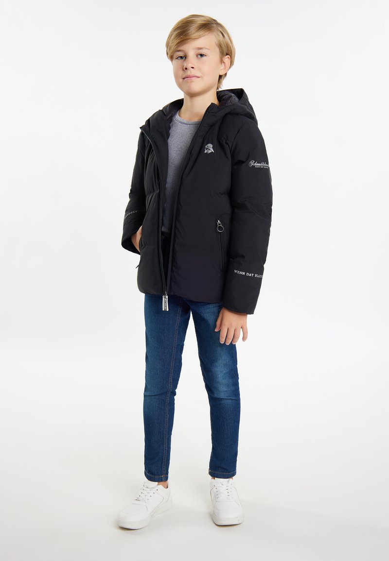 Svart puffjacka med dragkedja framtill, huva och broderade detaljer. Bärs över en grå skjorta, tillsammans med blå jeans och vita sneakers.