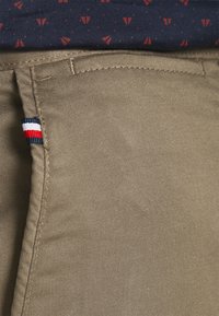 I pantaloni di cotone khaki presentano una texture liscia, un taglio dritto e una piccola etichetta a marchio blu, rossa e bianca sulla cucitura laterale.