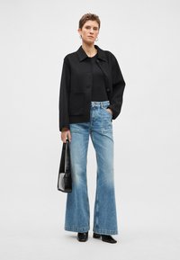 Casaco preto de mistura de lã com gola e bolso frontal, combinado com jeans azuis claros de perna larga e uma mala de pele preta.