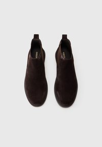 Une paire de bottes Chelsea en cuir suédé marron foncé, sans lacets, avec des panneaux élastiques sur les côtés et des languettes de traction, vue de dessus sur un fond blanc uni.