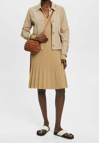 Beige kort jacka över en plisserad, knälång klänning i beige. Brun vävd handväska och vita sandaler med svarta sulor.