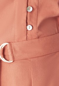 Tela color rosa con un cinturón que tiene una hebilla circular de plata, y dos botones redondos hechos de un material translúcido.