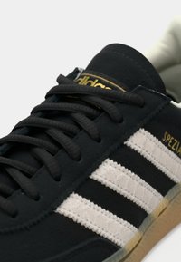 Μαύρο σουέτ αθλητικό παπούτσι με λευκές ραβδώσεις με υφή, με μαύρα κορδόνια και χρυσό λογότυπο. Στρογγυλεμένη μύτη και αντιθετική ελαστική σόλα.