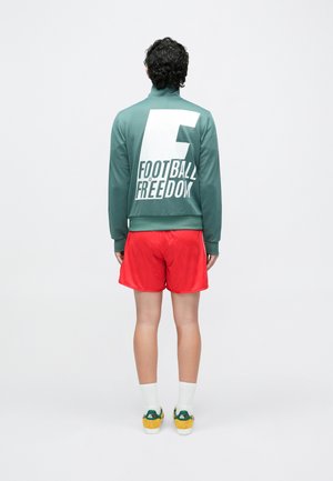 Personne debout de dos portant une veste verte avec le texte "Le football, c'est la liberté", un short rouge, des chaussettes blanches et des baskets jaune-vert.