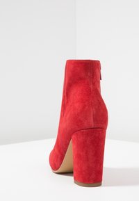 Bottines en daim rouge à bout arrondi et talon bloc haut. Elles sont munies d'une fermeture éclair à l'arrière et d'une semelle lisse couleur tan.