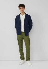 Marineblaue Jacke mit zwei Fronttaschen, weißes Poloshirt, olivgrüne Cargo-Hose mit Seitentaschen und weiße Sneaker.