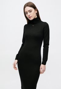 Νεαρή γυναίκα με ίσια καστανή τρίχα που φοράει εφαρμοστό μαύρο φόρεμα ζιβάγκο, όρθια μπροστά σε λευκό φόντο.