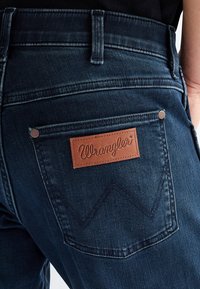 Povečava zadnjega žepa temno modrih kavbojk iz jeansa z rjavim usnjenim Wrangler logotipom in vidnimi šivi.