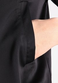 Black garment featuring a smooth fabric with a subtle texture. A hand reaches into a concealed side pocket, showcasing functional design elements.