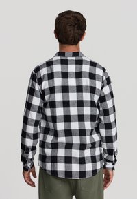 Uomo con capelli corti castani che indossa una camicia a maniche lunghe a scacchi bianco e nera e pantaloni verde oliva, girato di spalle.