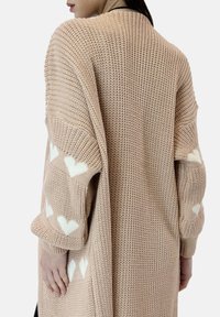Kardigan dziany w miękkim beżu z długimi rękawami ozdobionymi białymi wzorami serc. Luźny fason z teksturowaną tkaniną i swobodnym krojem.