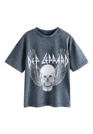 Person trägt weiße Hose, schwarz-weiße Sneakers und ein dunkelblaues T-Shirt mit weißem Totenkopf-Design, steht in der Nähe eines blau gepolsterten Stuhls.