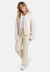 Beige gebreide zip-up jack met een brede kraag, gecombineerd met crèmekleurige broek en witte sneakers. De stof heeft een gestructureerde afwerking.