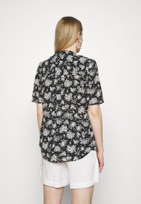 Chemise à manches courtes en tissu noir avec un motif floral blanc, avec un dos boutonné et un ourlet arrondi. Associée à un short en lin blanc.