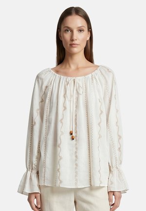 Jonge vrouw met steil haar draagt een losse, witte blouse met lange mouwen, beige borduursel en een gestrikte halslijn met houten kralen.