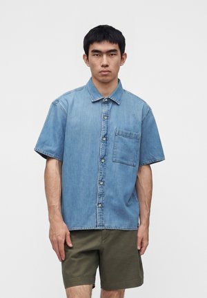Ung mand iført en kortærmet blå denimskjorte med brystlomme og olivengrønne shorts, der står foran en ensfarvet hvid baggrund.