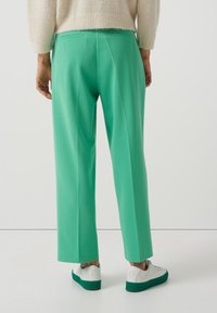 Gröna skräddarsydda byxor med rak passform och fickor, i kombination med vita sneakers med gröna sulor, mot en neutral bakgrund.