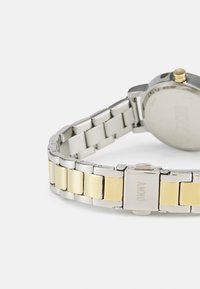 Ceas din metal cu tonuri de argint și aur cu o brățară din metal în două tonuri. Dispune de o carcasă rotundă și un dispozitiv de închidere cu buton. Finisaj metalic, fără modele.