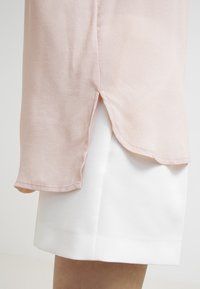 Blouse rose clair en tissu texturé et transparent avec une fente sur le côté, associée à une jupe blanche structurée. La blouse a un ourlet arrondi.