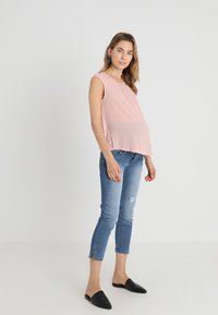 Top de maternidade rosa sem mangas com um corte confortável, combinado com jeans skinny despedaçados de um azul claro e mules pretos sem fecho.