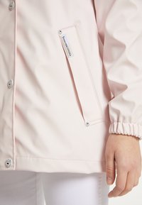 Veste imperméable rose clair avec boutons-pression, poignets élastiques et une poche latérale ornée d'une étiquette logo. Texture lisse, finition légèrement brillante.