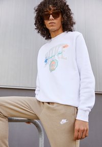 Biely sweatshirtn so multicolor grafickým dizajnom značky Nike a béžovými teplákmi s malým bielym logom Nike. Model nosí slnečné okuliare a má kučeravé vlasy.