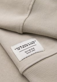 Tessuto a coste beige con un'etichetta iscritta "L'UNICO TUO LIMITE È LA TUA MENTE," con il nome del marchio "OMBRE" e l'anno di fondazione "dal 1985."