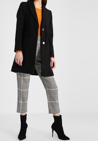 Frau mit schwarzem Knopfmantel, orangefarbenem Oberteil, karierten, verkürzten Hosen und schwarzen, hochhackigen Stiefeletten vor einem weißen Hintergrund.