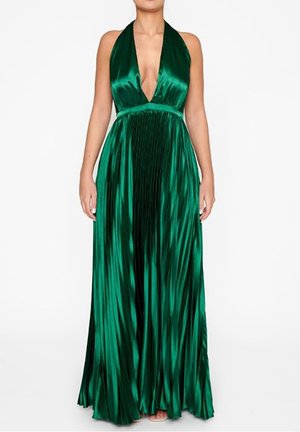 Femme portant une robe de soirée plissée vert émeraude brillante, sans manches, à décolleté en V profond, debout avec les bras relâchés.