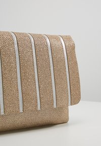 Złota kopertówka z brokatem o pionowym paskowanym wzorze i srebrnych akcentach. Teksturowana powierzchnia, klapka złożona do środka. Kompaktowy, prostokątny kształt.