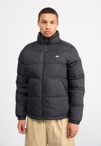 Giacca a puffo nera isolata con colletto alto, chiusura con zip e polsini elasticizzati; presenta motivi a righe orizzontali e dettagli con logo.
