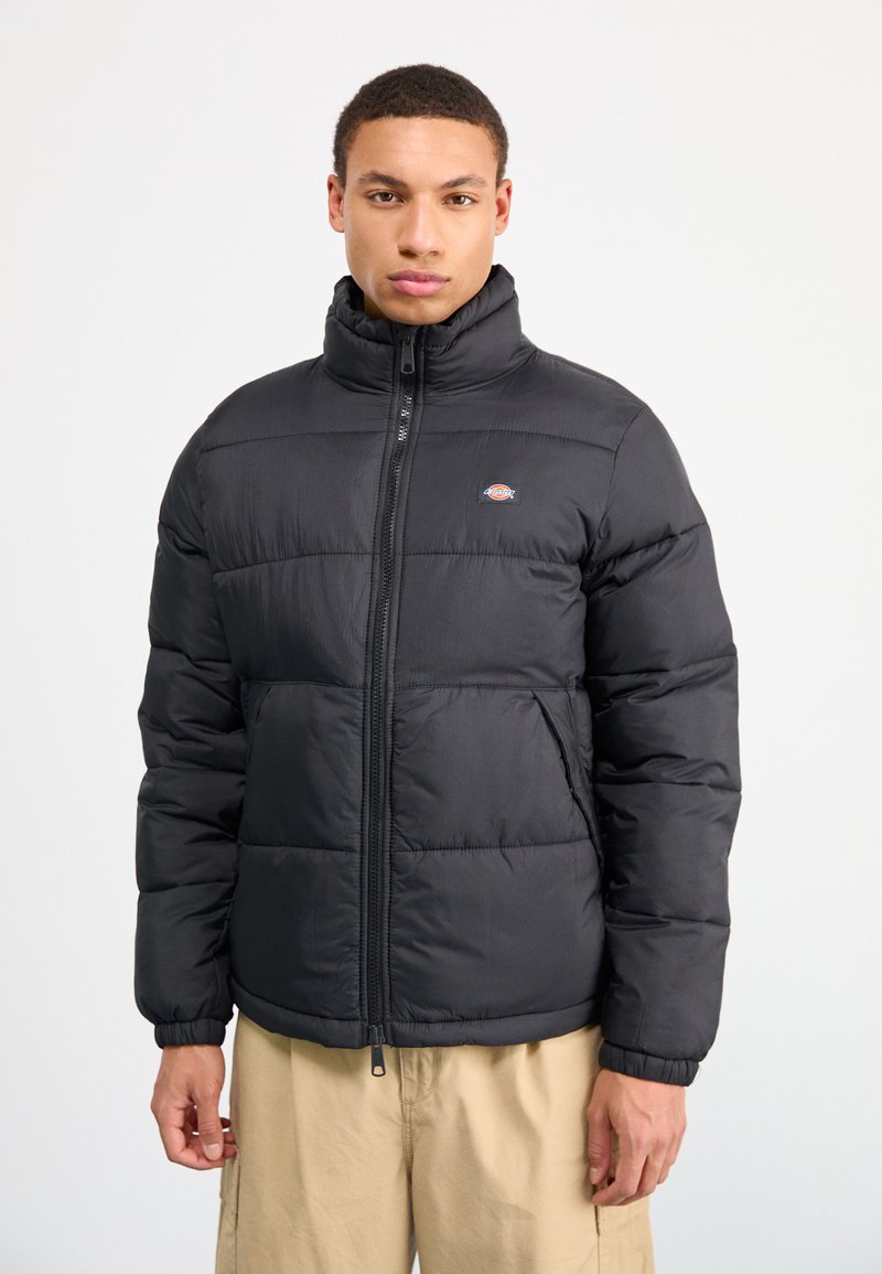 Giacca a puffo nera isolata con colletto alto, chiusura con zip e polsini elasticizzati; presenta motivi a righe orizzontali e dettagli con logo.