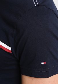 Námořnické modré tričko s krátkým rukávem, vyrobené z bavlny. Na rameni má červený a bílý pruh a na rukávu malé logo značky.