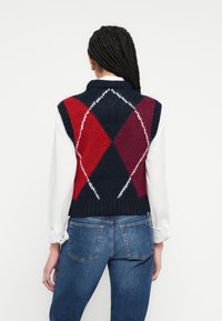 Pletená vesta s tmavomodrým podkladom a červeným a vínovým kosoštvorcovým vzorom. Lem a golier sú rebrované, doplnené o biele tričko s dlhým rukávom.