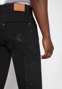 Czarne spodnie cargo z denimu o teksturowanej tkaninie, z haftowanym logo i skórzaną łatą na talii. Posiada liczne kieszenie i nitowane detale.