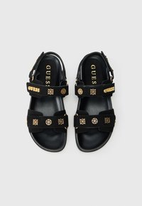 Μαύρα σανδάλια από σουέντ με χρυσές λεπτομέρειες, διαθέτοντας φαρδιές λουρίδες και διακοσμητικό εξάρτημα. Η εσωτερική σόλα είναι μαύρη, με τη σήμανση "GUESS".