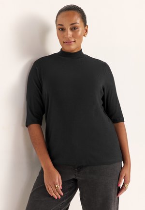 Schwarzes kurzärmeliges Rollkragenoberteil mit geripptem Stoff, lockerem Schnitt und geschwungenem Saum. Kombiniert mit dunkelgrauer Hose. Einfaches und vielseitiges Design.